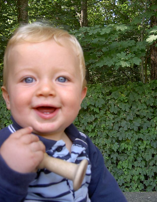 Oskar Vogel mit Holzspielzeug in der Hand vor grün bewachsenem Zaun.